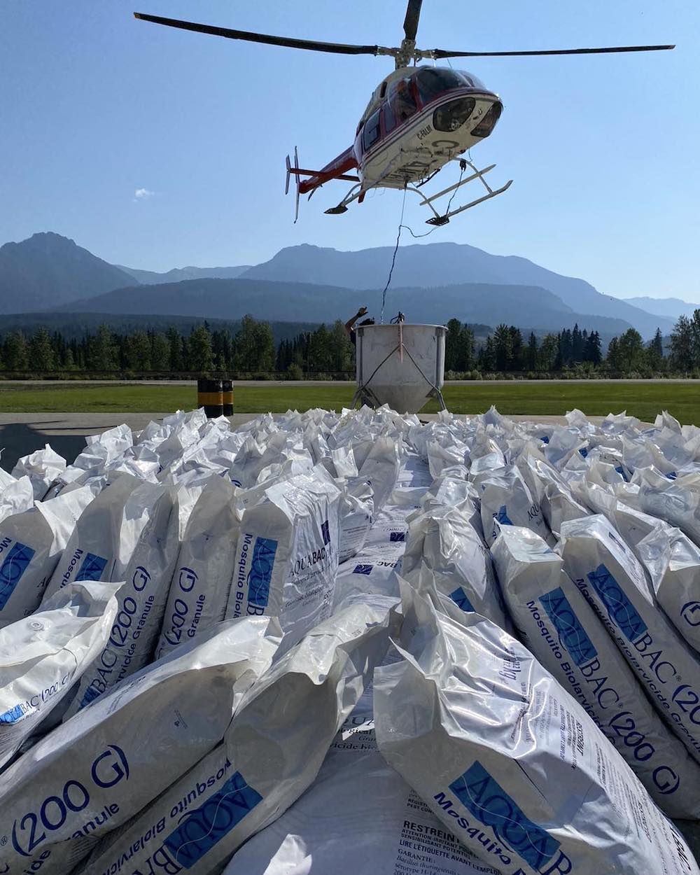 A white and blue helicopter flies low over a wide collection of white and blue bags marked '200 g.' In the background is a green farmer’s field and a blue mountain range.