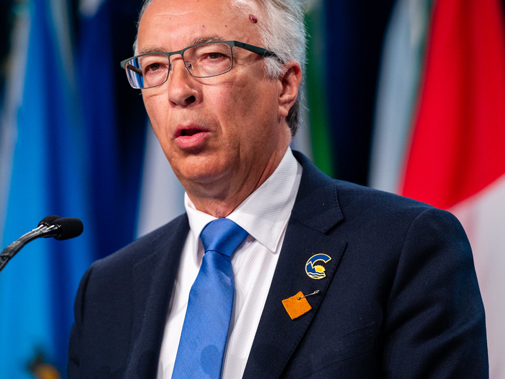 A 60-ish light-skinned man in a suit and blue tie stands at a podium. He is surrounded by flags.