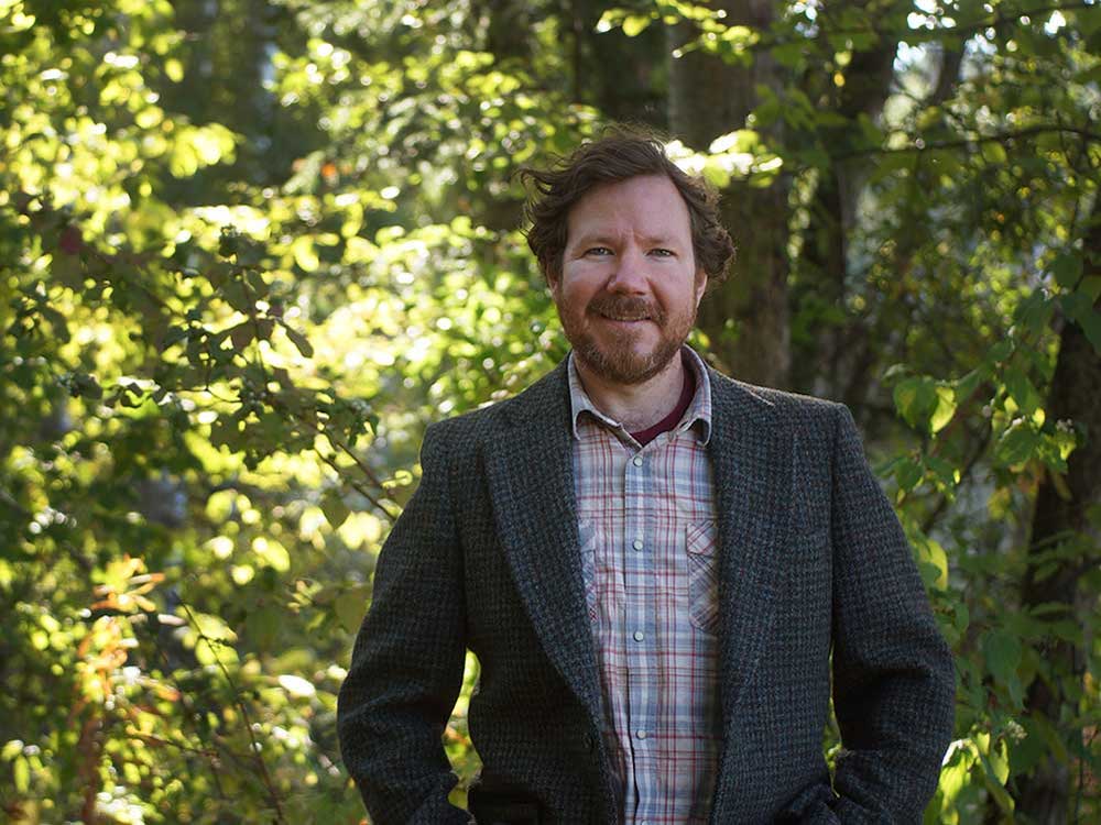 A man with short brown hair and a brown beard, wearing a plaid shirt and suit jacket, looks at the camera. He stands in front of a forested background.