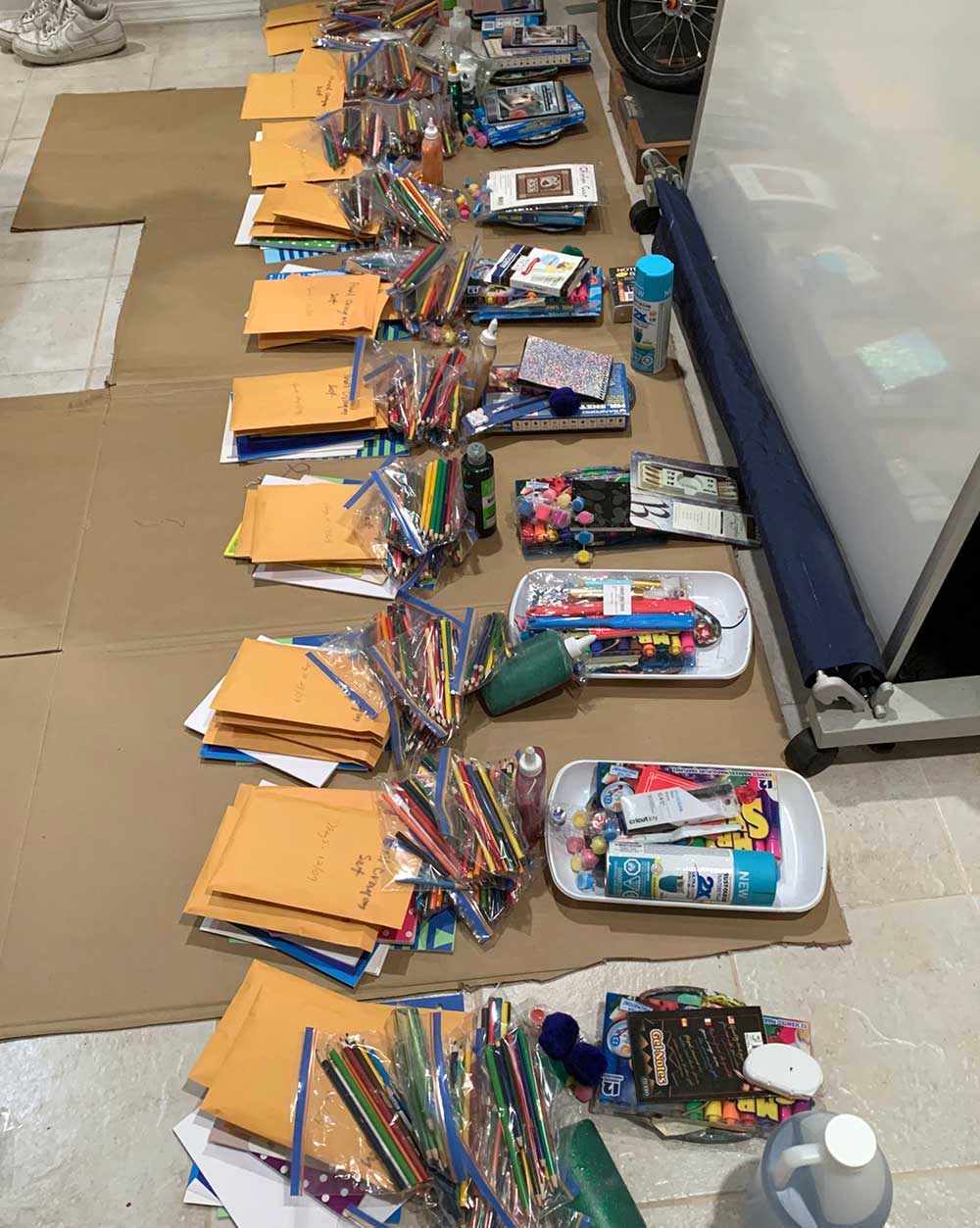 Ten assembled packs of paints, paper, coloured pencils and paint trays are lined up on a tiled floor.