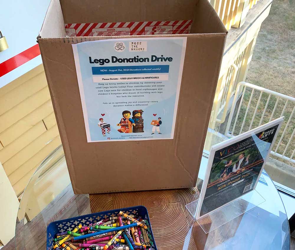 A medium-sized cardboard box sits on a table outside a yellow building. On the box is a light blue poster asking for Lego donations. A green plastic basket of crayons sits on the table in front of the box.
