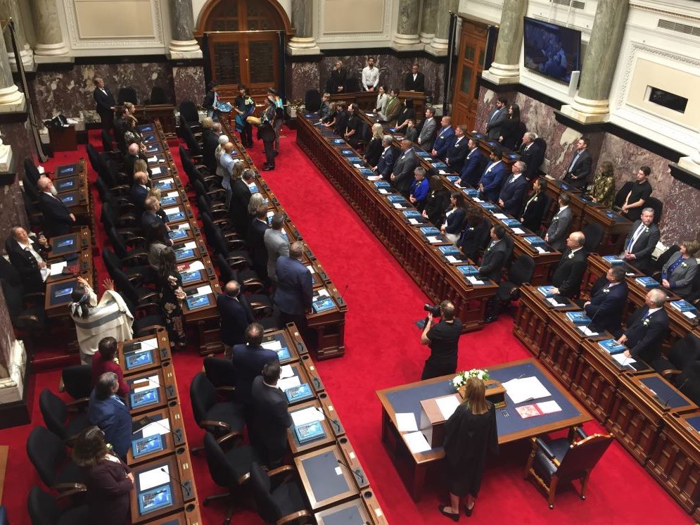 View from above of a room full of people wearing suits standing at desks in the B.C. legislature.