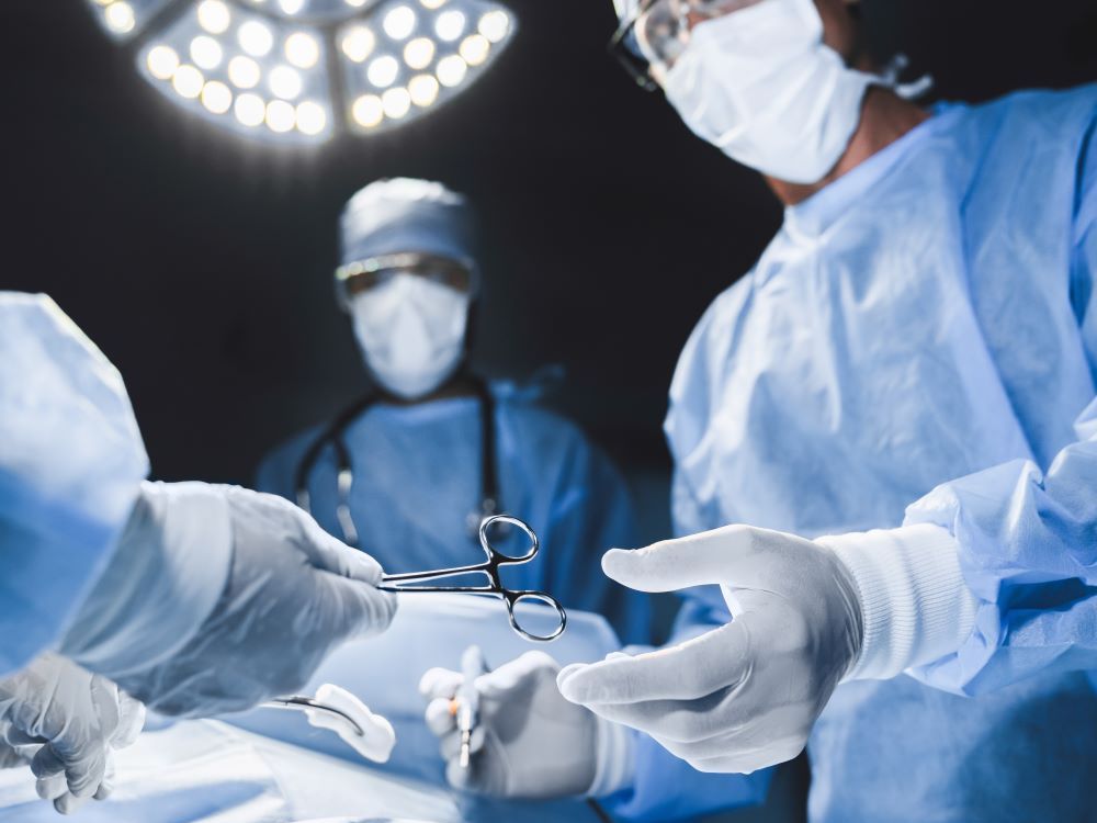 Under the bright lights of an operating room, a white-gloved surgeon reaches for a pair of scissors being handed to them by someone out of the frame.