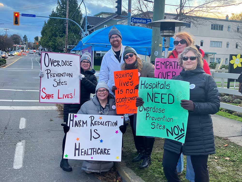 Six people stand on a grass strip near the road holding protest signs. The blue tent of the OPS is visible in the background.