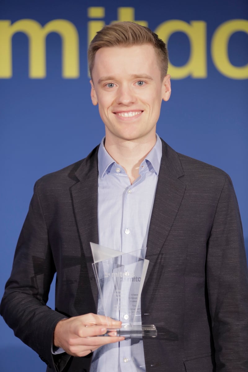 A young man with light skin tone and short light hair wears a suit and holds a clear glass award.