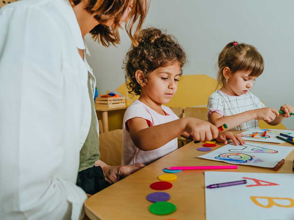 A kindergarten-level English lesson. Students are colouring the letter A, accompanied by a teacher.