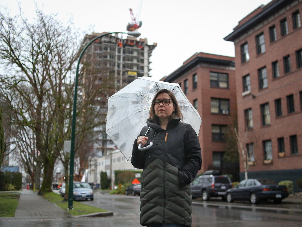 Teresa Alfeld stands in a dark green and black puffy winter jacket. She has short dark hair and glasses with thick frames. She holds a transparent umbrella and is standing on the corner of a residential street on a grey day. A red brick four-storey walk-up apartment is behind her, and in the background is a highrise apartment building under construction.
