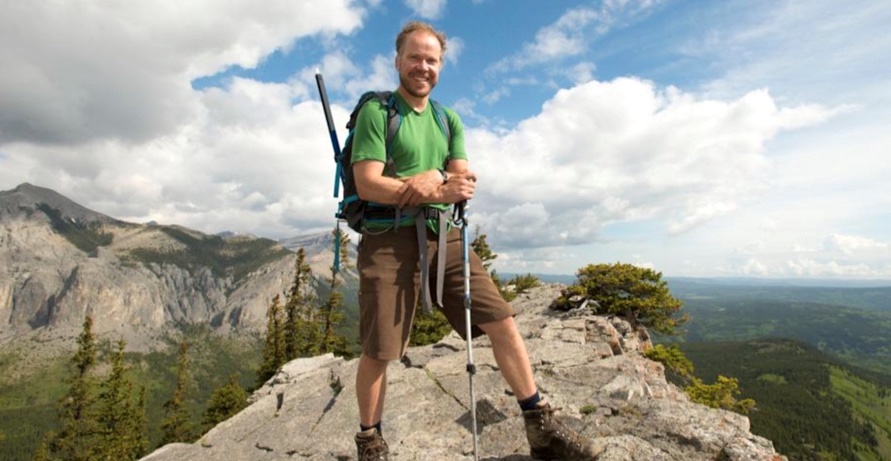 A man with light skin and light brown hair stands on a rocky ridge with views of forests below and mountains behind. He is wearing a green T-shirt, brown shorts and hiking boots and holds a walking stick in front of him.