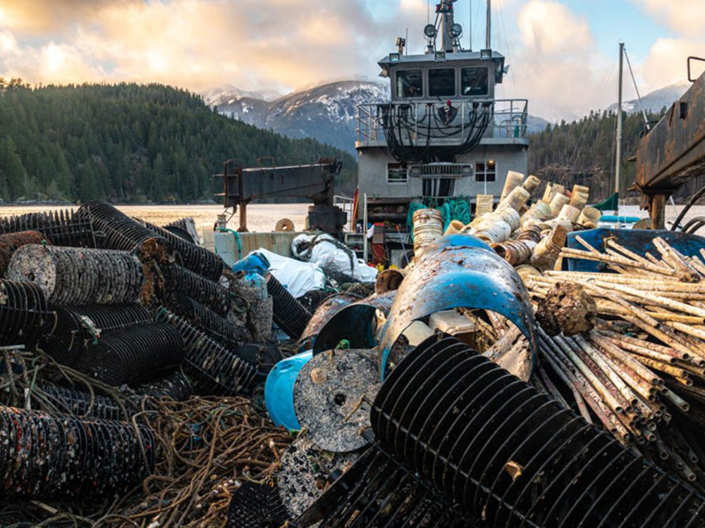 A vessel with a flat bed and crane is full of ocean debris such as PVC pipes, blue plastic barrels, white plastic tubs and plastic shellfish baskets.