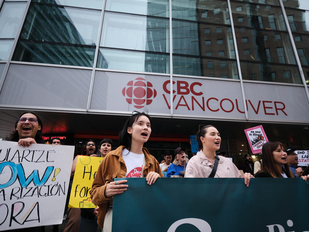 People protest outside the CBC building in Vancouver. They are holding signs saying things like 'No One Is Illegal. Canada Is Illegal.'