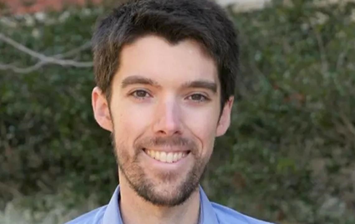 A headshot of a young man with light skin, short dark brown hair and wispy beard.