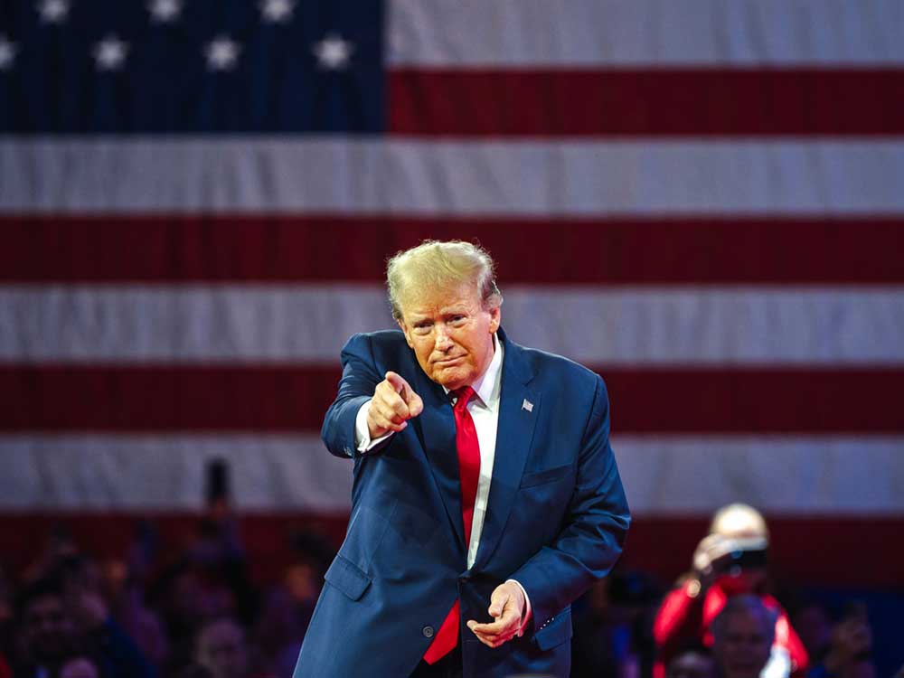 Donald Trump wears a dark blue suit, white shirt and red tie. There is a giant U.S. flag behind him. He points at the crowd.
