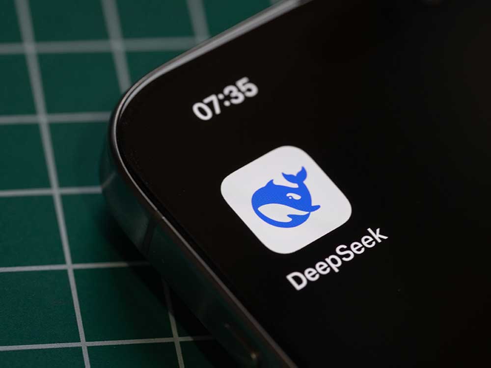 A smartphone sits on a desk. On the screen is the DeepSeek app, with its logo of a stylized whale.