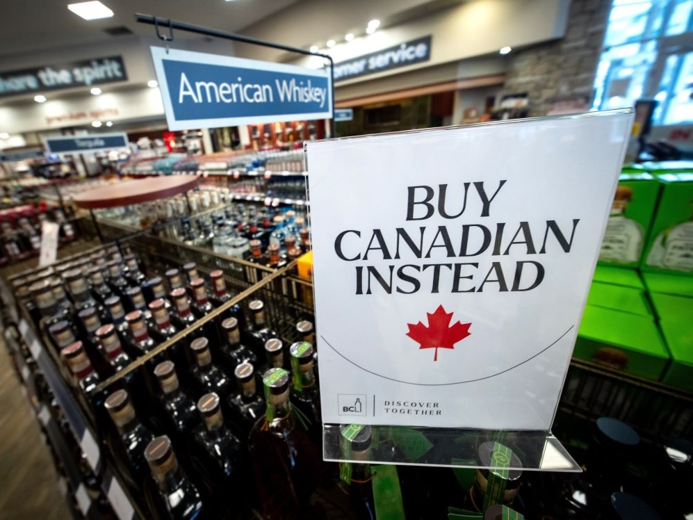 In the American whiskey section of a liquor store, a sign placed on top of a shelf of bottles reads 'Buy Canadian instead' with a red maple leaf.