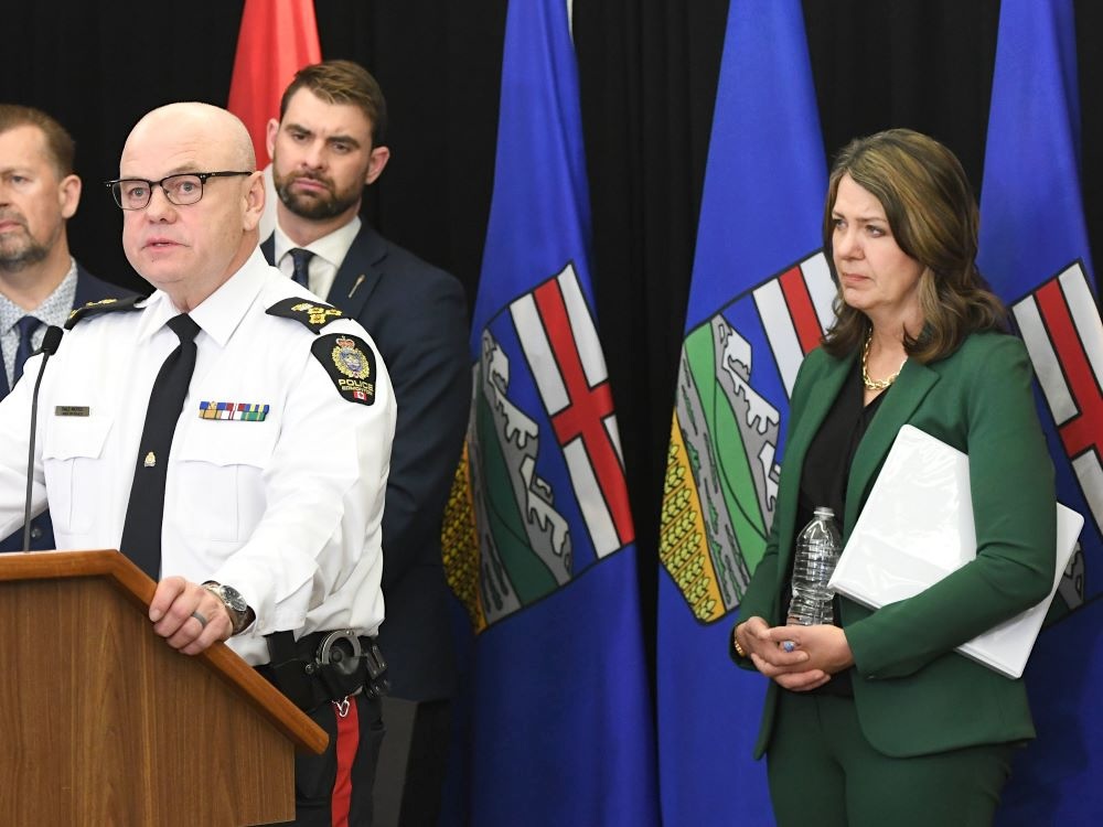 At left, a light-skinned bald man in a white and black police uniform speaks into a microphone at a podium. At right, a light-skinned woman with shoulder-length brown hair looks on. She is wearing a green pant suit, has a white binder under one arm and holds an open plastic water bottle. Several Alberta flags are behind them.