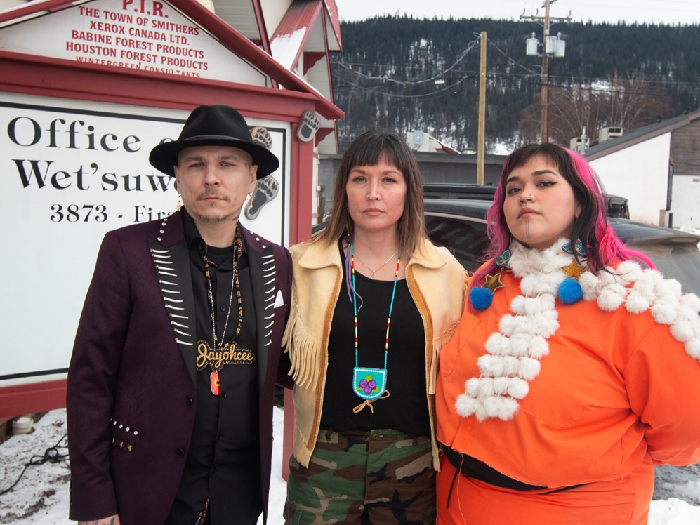 Three people, one man and two women, stand in an outdoor setting next to a sign that says 'Office of the Wet’suwet’en.'