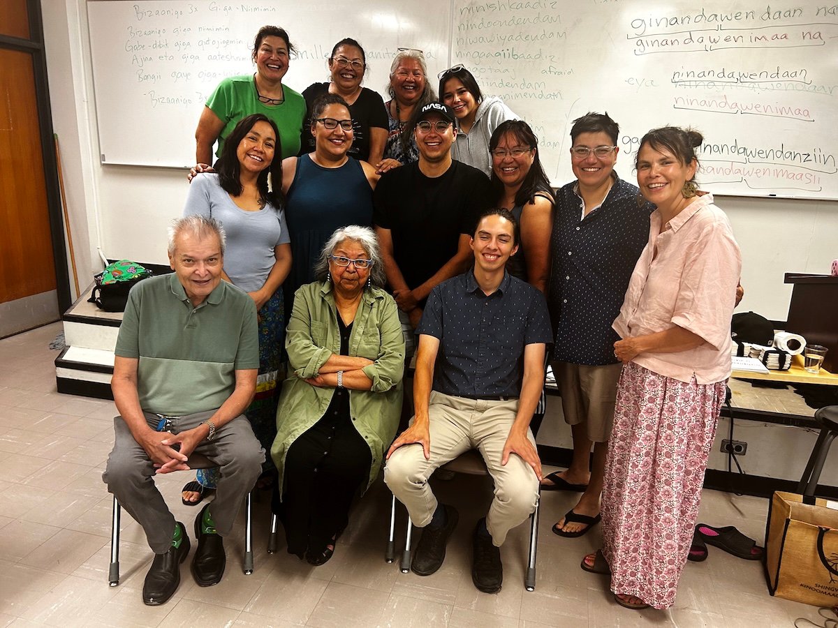 Thirteen people pose for a photo in a classroom. They are all smiling, and a range of different ages. Behind them, there are Ojibwe phrases written on the whiteboard.