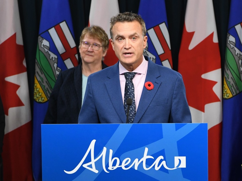 A light-skinned man with short grey-brown hair, wearing a blue jacket with a poppy pin, a pink shirt and navy patterned tie, speaks from behind a podium that says 'Alberta.'