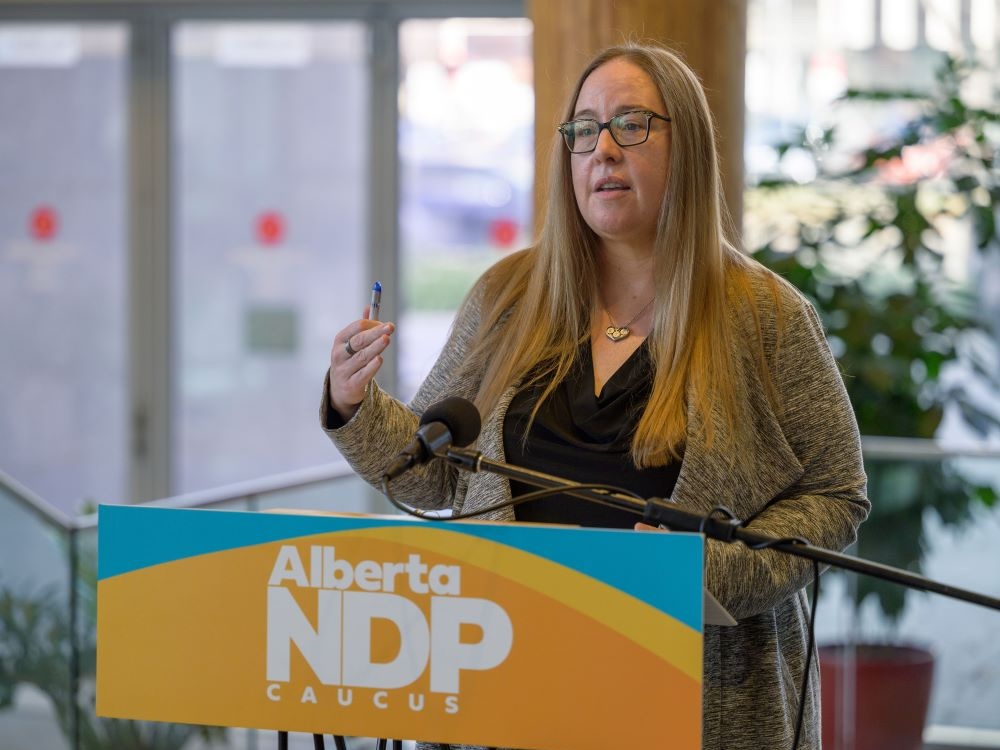 A woman with light skin, long straight blond hair and glasses stands at a podium that reads 'Alberta NDP caucus.' She is gesturing with one hand and appears to be mid-speech.