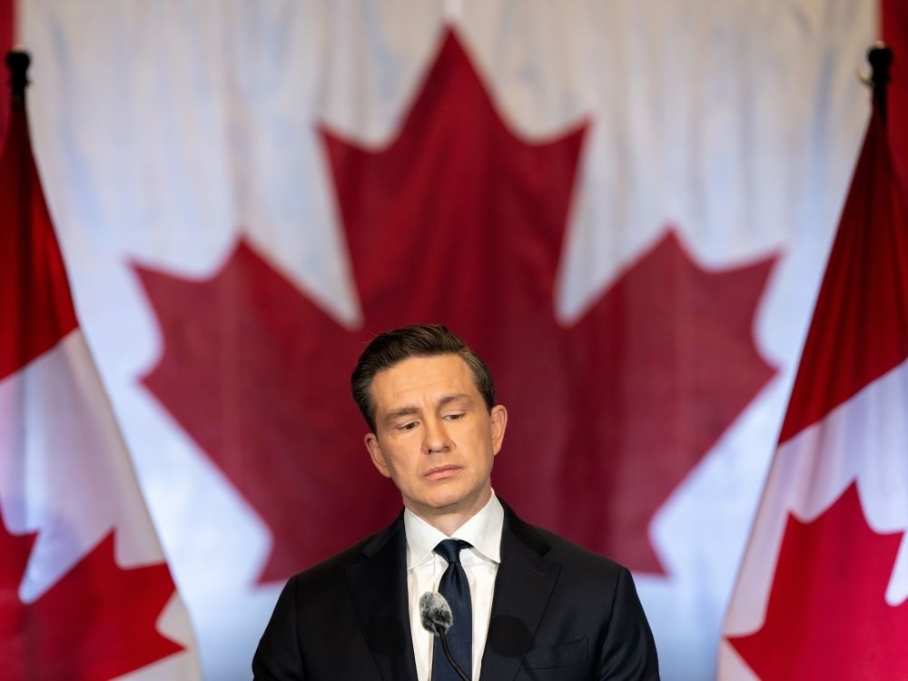 An unsmiling Pierre Poilievre, a light-skinned man with short dark hair wearing a black suit, looks downward behind a microphone. His head is centred in front of the large red maple leaf of a Canadian flag behind him.