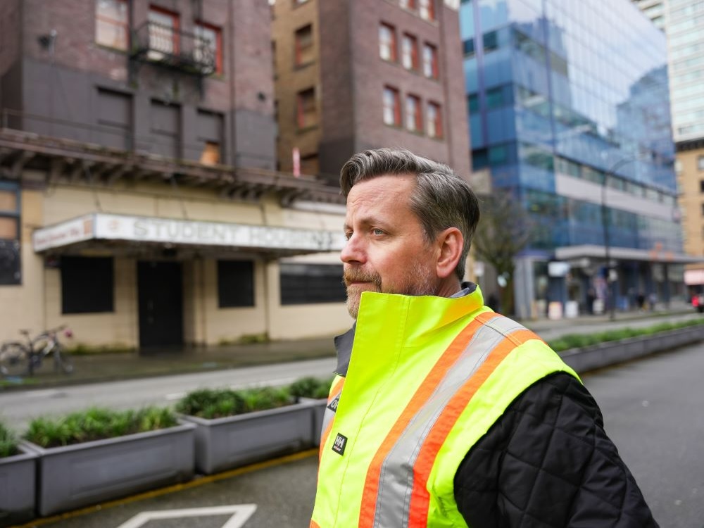 A middle-aged man with light skin tone and salt-and-pepper hair wears a hi-vis vest and a black jacket. In the background, across the street, is Dunsmuir House.