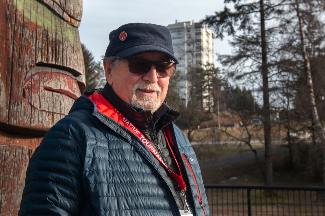 A man wearing a jacket, cap and sunglasses stands in an outdoor setting. There is a totem pole behind him.