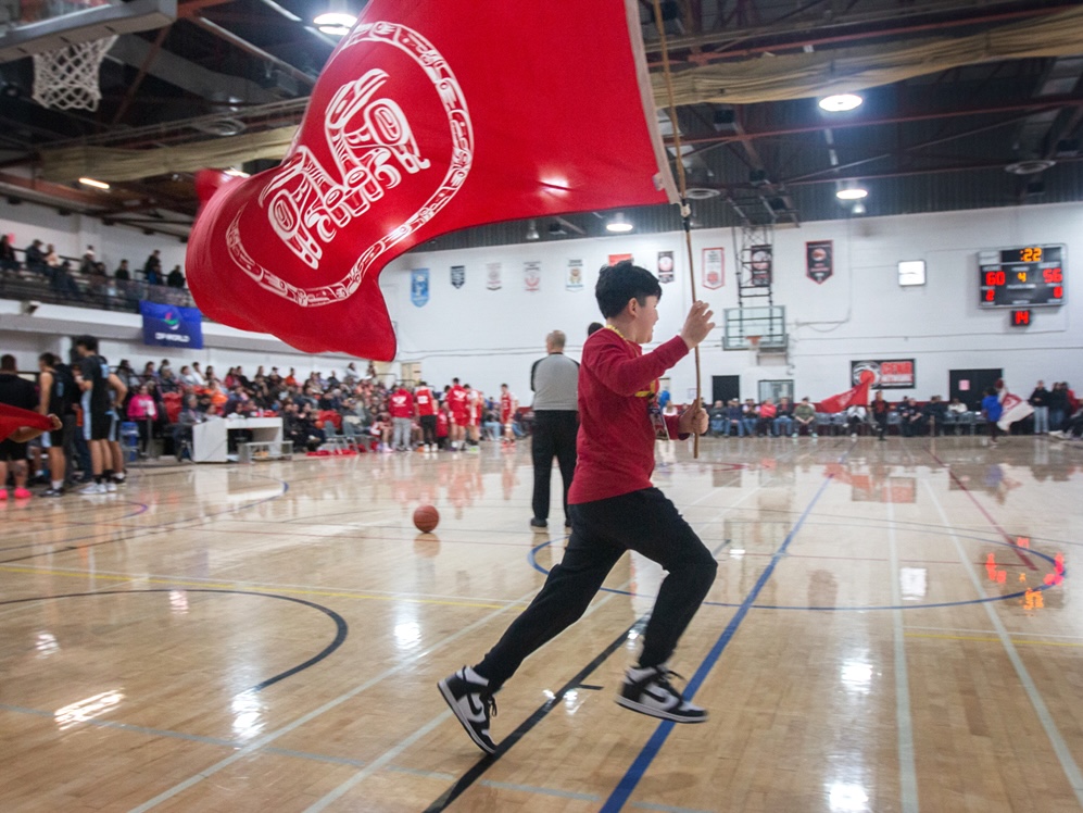 A child runs through a basketball court carrying a red flag that bears an Indigenous design. There are players and fans in the background.