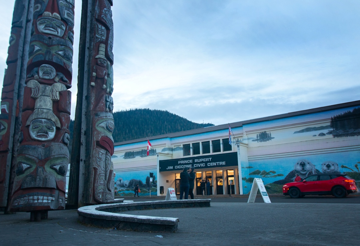 People gather in front of a large building with a sign over the door that says 'Prince Rupert Jim Ciccone Civic Centre.' There is a totem pole and benches in the foreground.