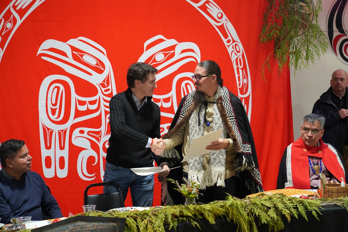 Two men stand behind a table holding sheets of paper and shaking hands. The man on the left wears a sweater and jeans while the man on the right wears glasses, his hair in a braid and traditional Indigenous regalia. Behind them is a red banner with an Indigenous-style insignia.