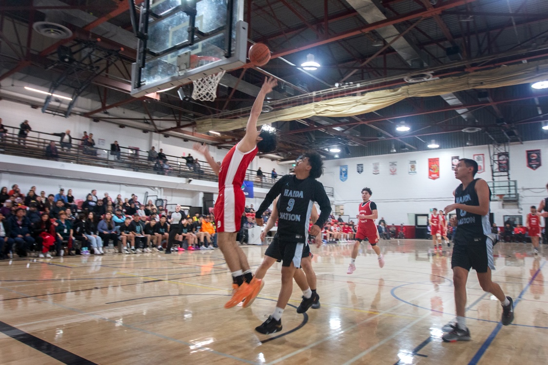 A player wearing a red Lax Kw'alaams uniform goes for a rebound, trailed closely by a player in a black Haida uniform.