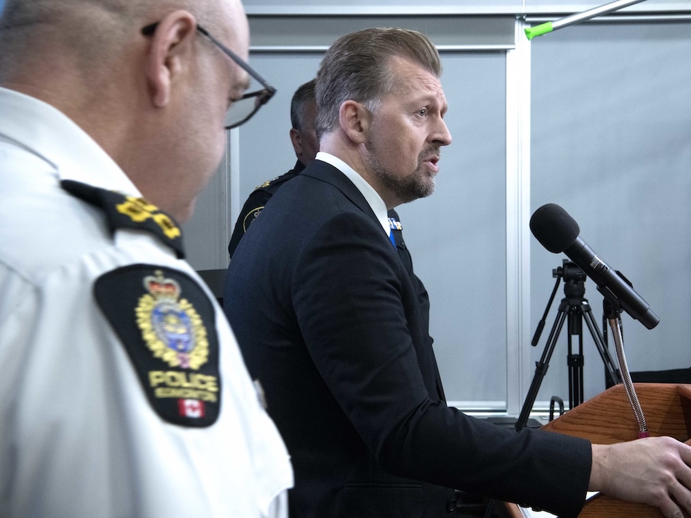 Side view of a man with light skin and light brown hair and beard, wearing a dark suit, standing at a podium speaking into a microphone. The profile of a bald man wearing an Edmonton police uniform can be seen to the left of the frame.