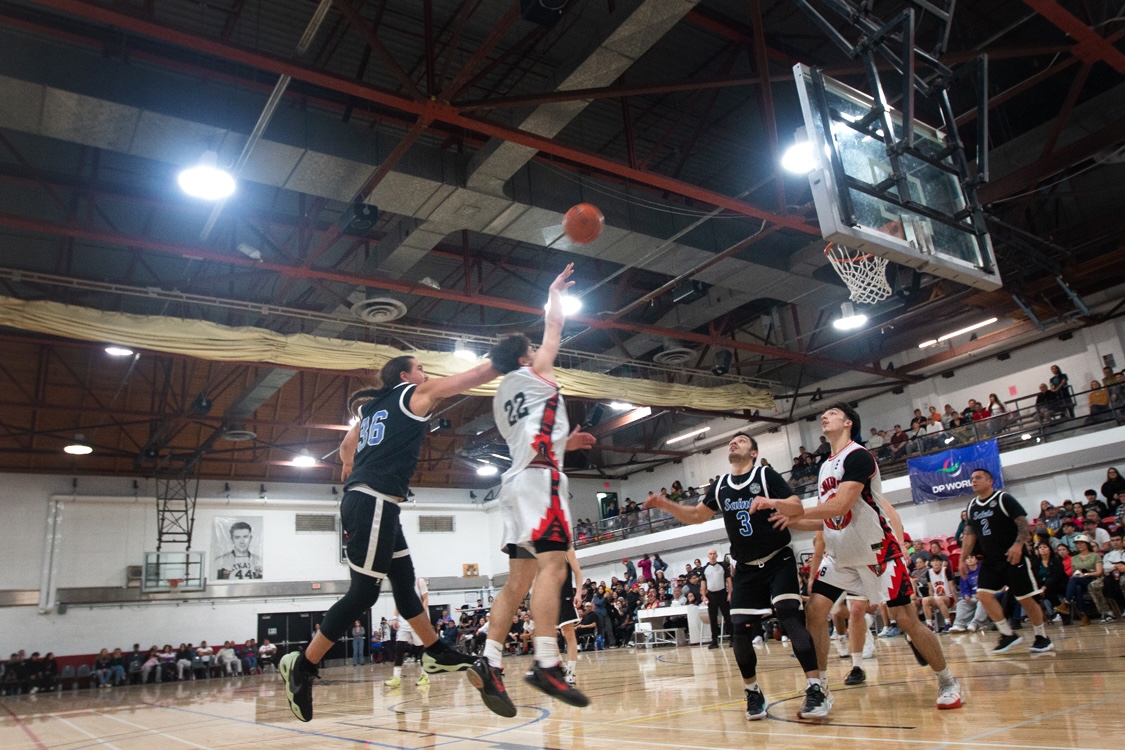 Two players leap for a ball in the middle of a basketball court. Two additional players stand opposite them, looking up at the ball. The stands surrounding the court are filled with people.