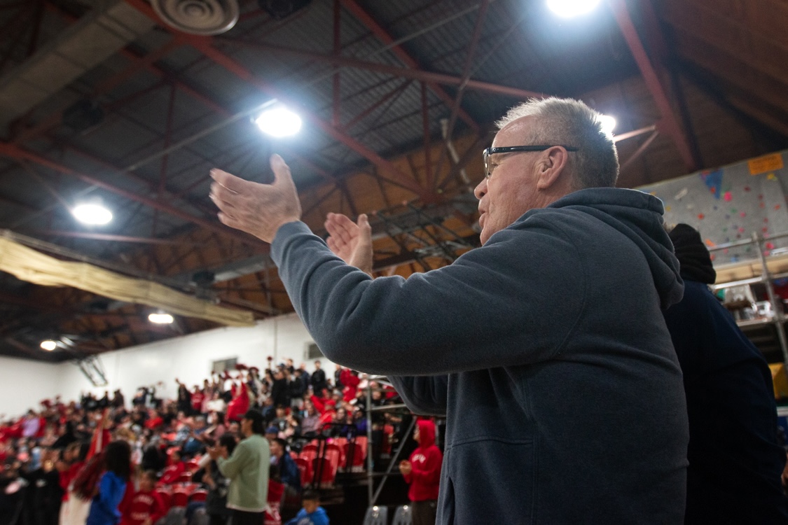 A man with short grey hair, wearing glasses and a navy blue hoodie, stands and claps. Behind him are stands filled with people.