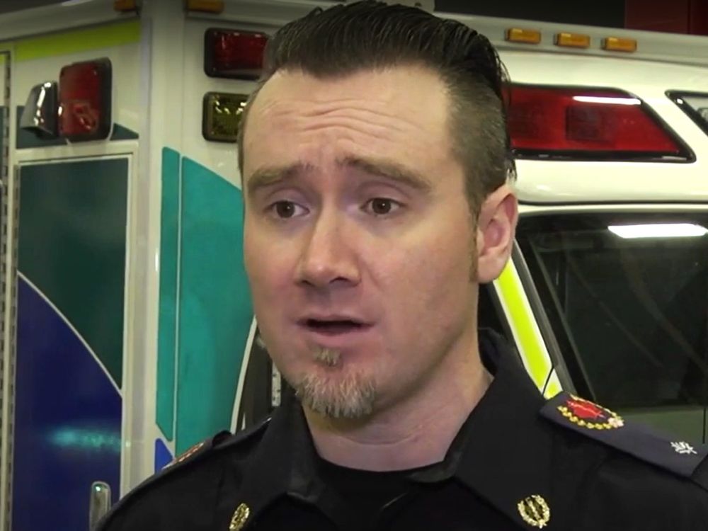 A light-skinned man with slicked back hair and a goatee and in a paramedic uniform speaks to a camera, with an ambulance behind him.