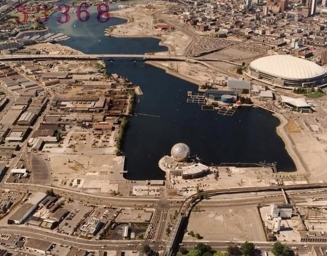A sepia-toned aerial view of False Creek in 1988, with the white roof of BC Place stadium to the right and the geodesic dome of Science World in the foreground, surrounded by empty lots and low-rise industrial buildings.