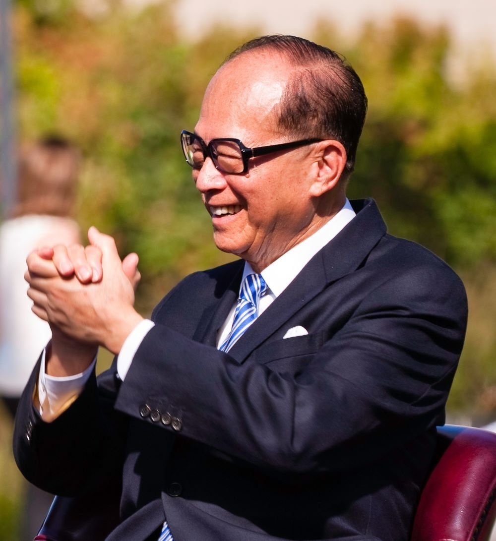 Li Ka-shing, a man with medium-light skin, thinning black hair and black thick-framed glasses wearing a navy suit, smiles widely and clasps his hands in front of him.