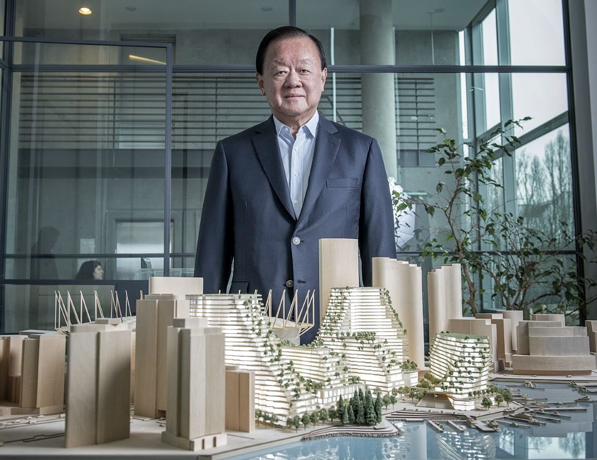 Oei Hong Leong has medium-light skin and short black hair and is wearing a navy blue suit over a white button-down shirt. He is standing in a glass-paned office behind an elaborate model of the Plaza of Nations redevelopment.