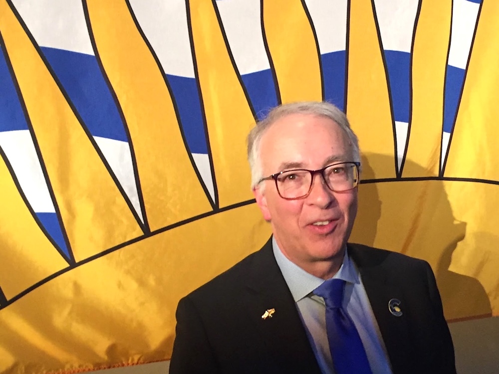 A 61-year-old light-skinned man with grey hair and glasses, wearing a suit and tie, poses in front of a large B.C. flag.
