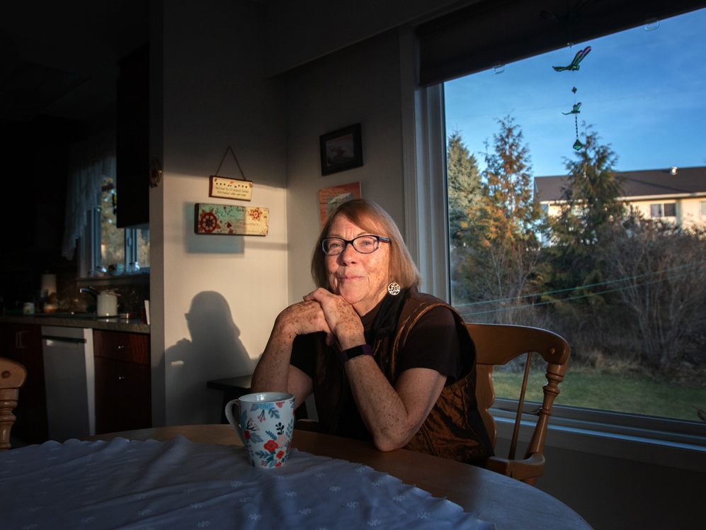 A woman wearing glasses sits at a table with her hands folded under her chin. There is a window behind her and a mug in front of her on the table.