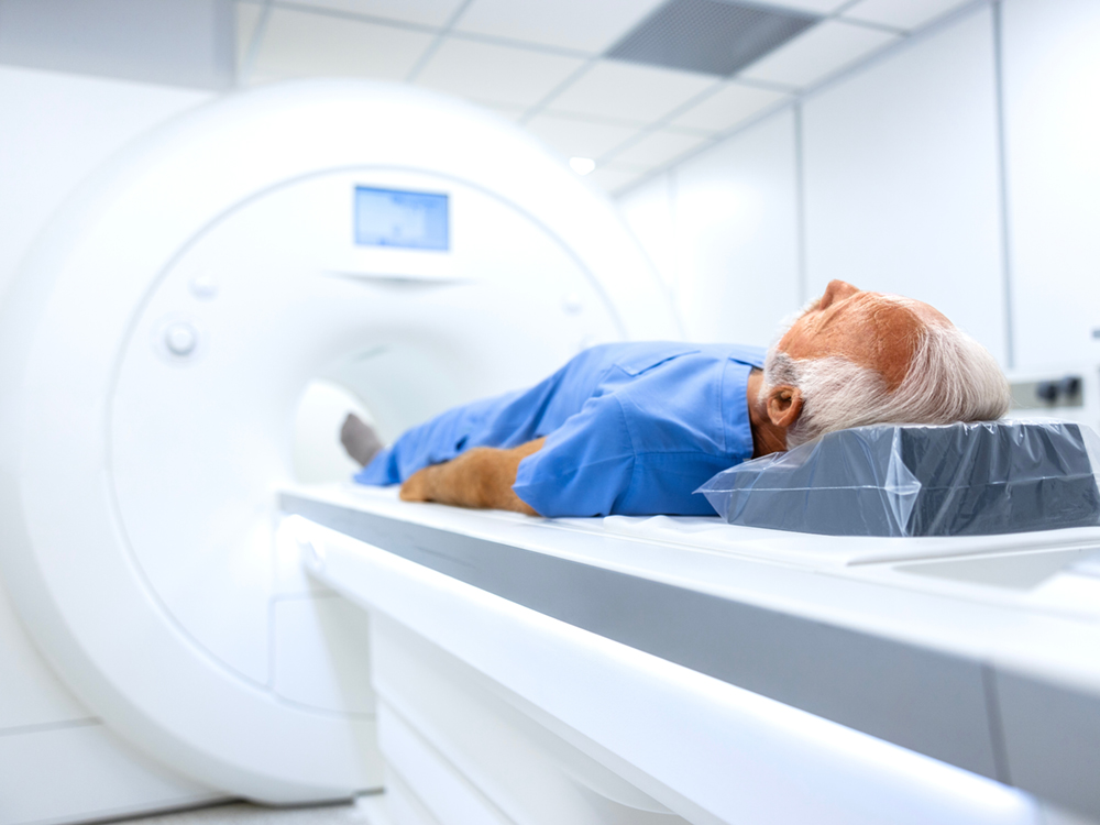 A person with a white, receding hairline gets ready for an MRI. They’re wearing a blue medical robe and their feet are pointed towards the MRI machine.