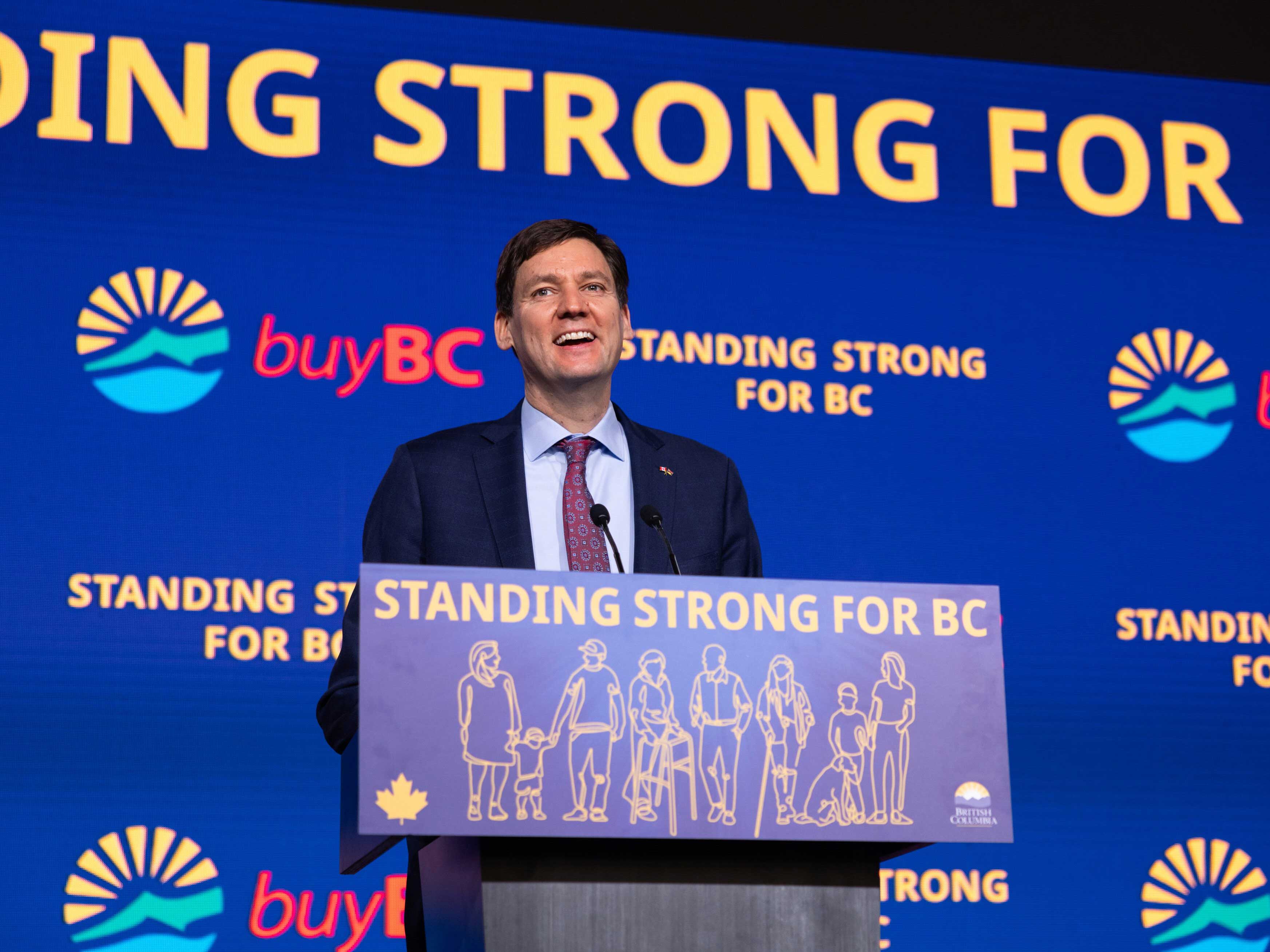 A 48-year old man in a dark suit stands at a podium in front of a blue backdrop that says 'Standing Strong for BC' and 'Buy BC.'