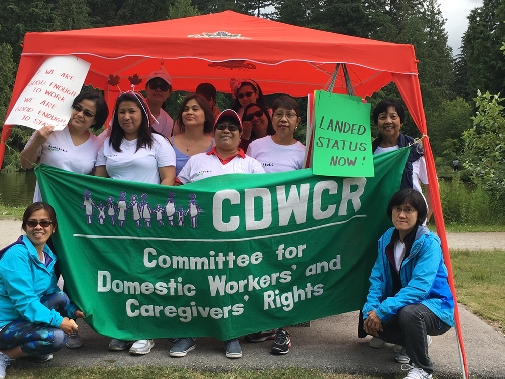 People from the Vancouver Committee for Domestic Workers&rsquo; and Caregivers&rsquo; Rights meet under a tent, holding a banner and signs. The banner is emblazoned with the committee name. A sign reads, &lsquo;If we are good enough to work, we are good enough to stay.&rsquo;
