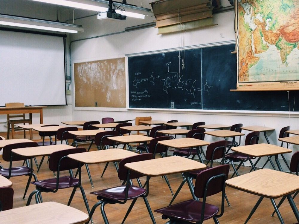 An empty classroom full of desks. A projector screen is pulled down at the front of the classroom. The side of the classroom shows some chemistry teaching chalked out on a chalkboard. There is also a world map.