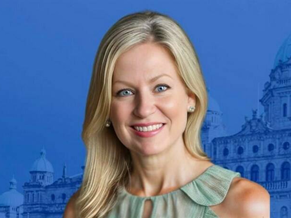 A 40-ish light-skinned woman with blond hair and a light-green shirt smiles at the camera. An image of the B.C. legislature is behind her.