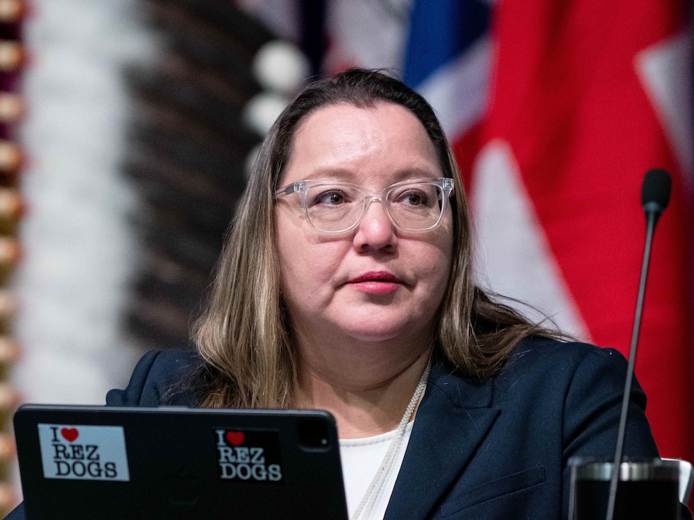 Cindy Woodhouse, a 40-ish Indigenous woman with a shoulder-length brown hair and glasses with clear frames, sits behind a microphone.