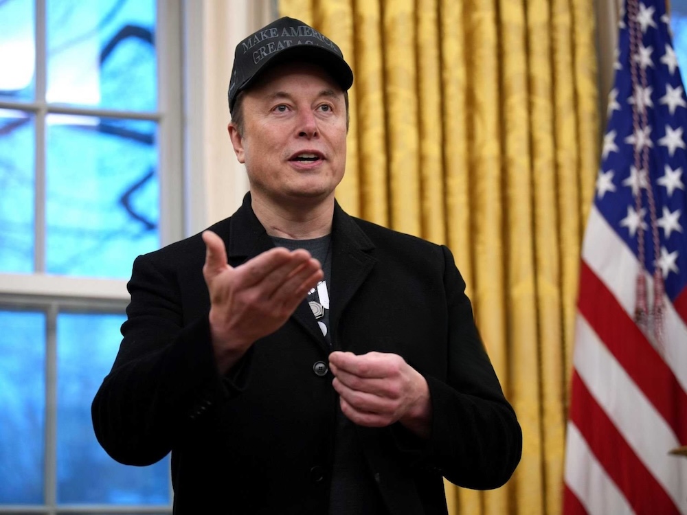 Elon Musk, a 53-year-old light-skinned man, is in the Oval Office wearing a black T-shirt and jacket and ball cap.