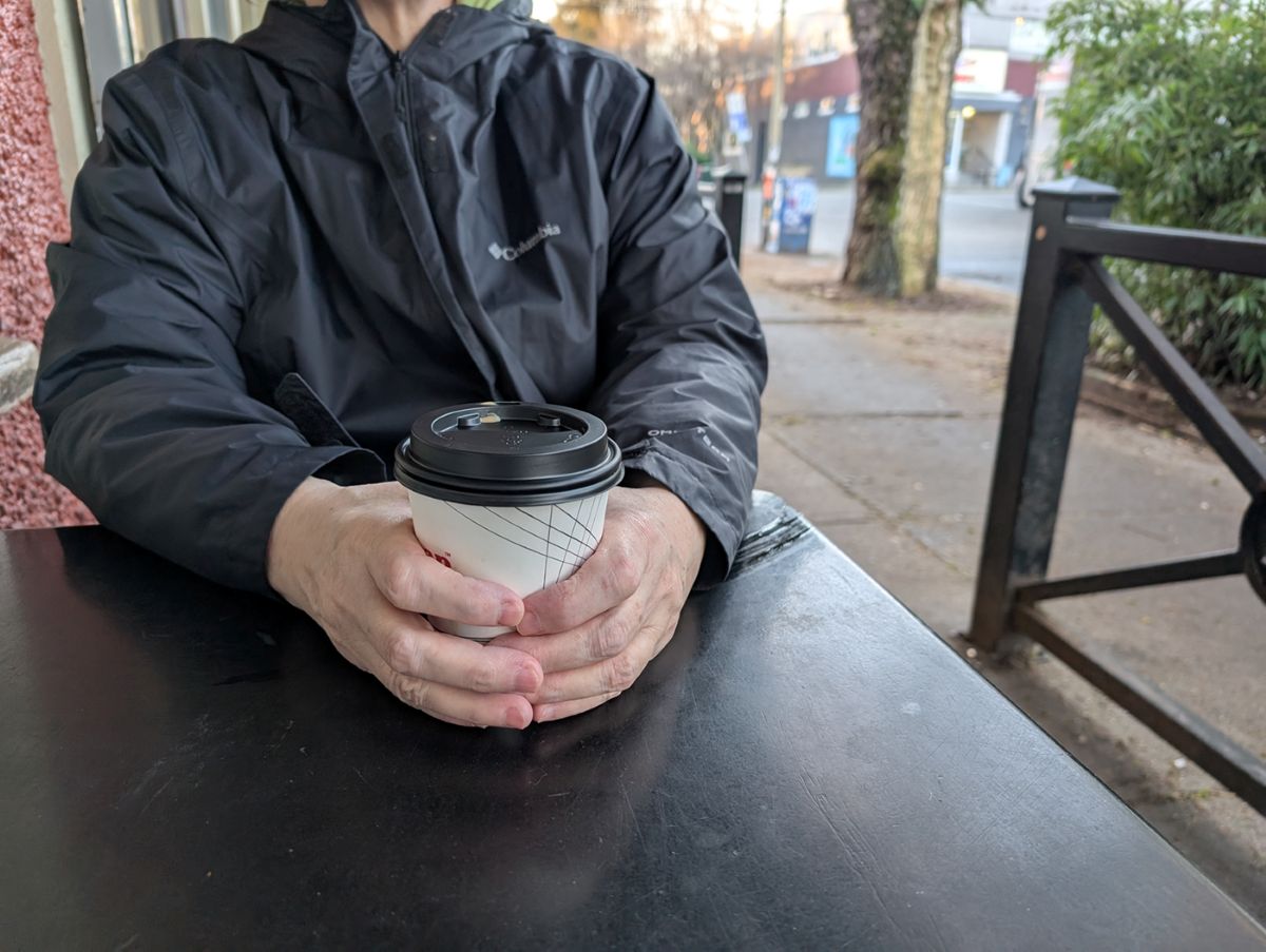 A close-up of a woman’s hands. She is sitting outside, holding a JJ Bean takeout coffee cup.