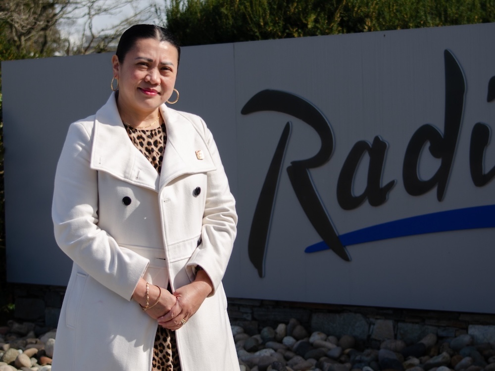 A woman with medium-toned skin wearing a white coat smiles in front of a hotel sign.