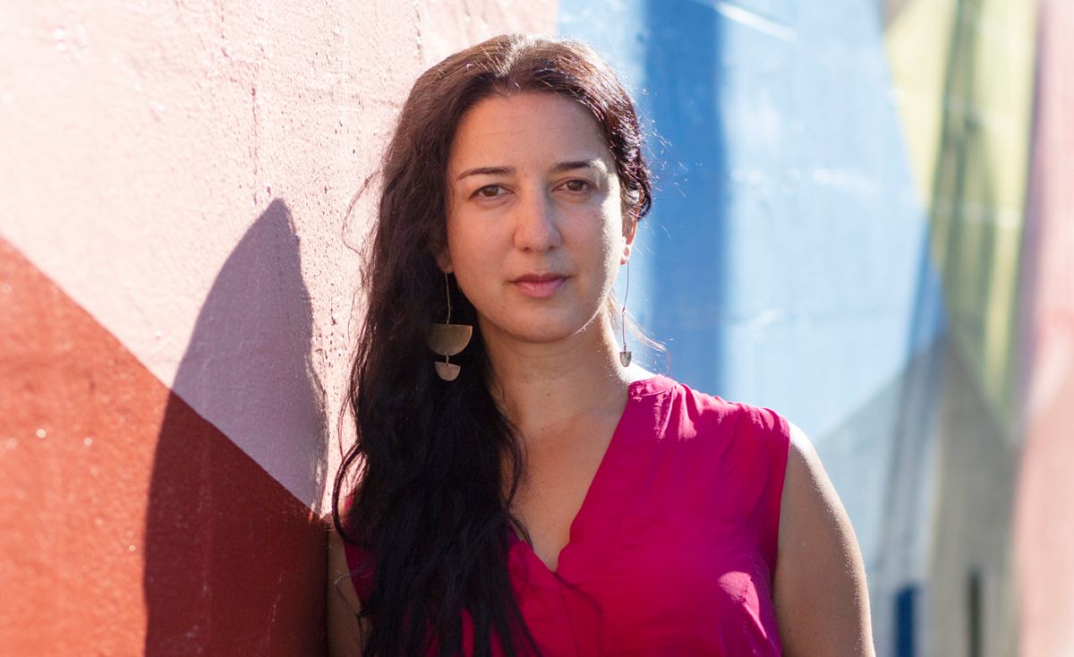 Kasari Govender, a woman with light skin tone and long wavy dark hair, stands next to a mural-painted wall outdoors. She is wearing a pink sleeveless shirt.