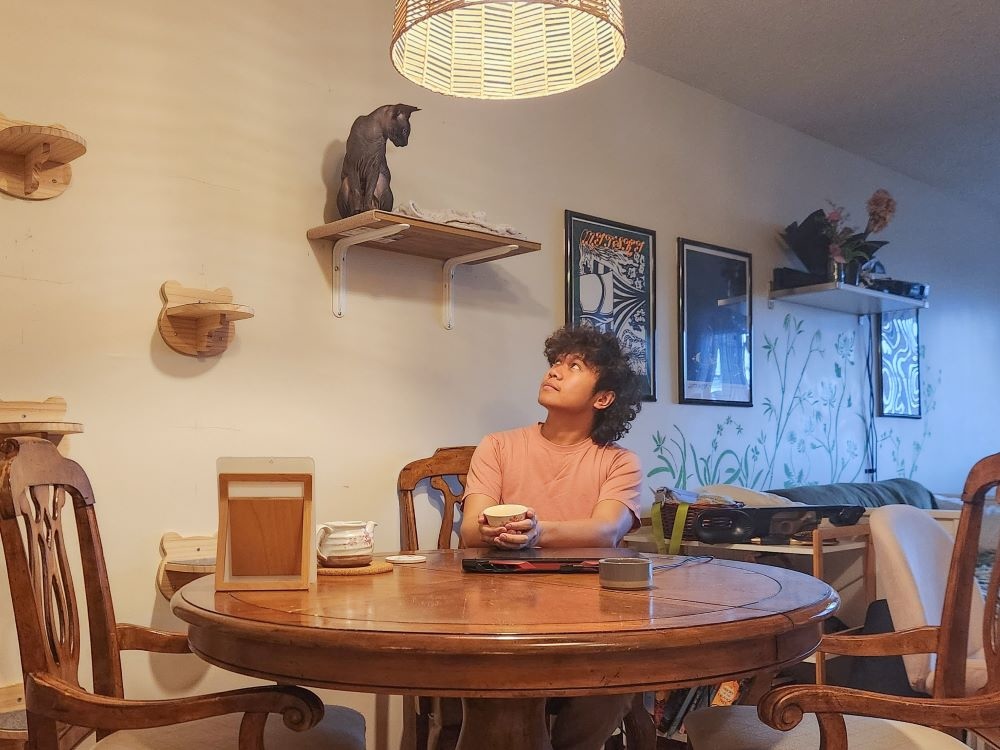 A man sits at a table inside an apartment, holding a cup of coffee and looking up at a hairless cat on the shelf just above him.
